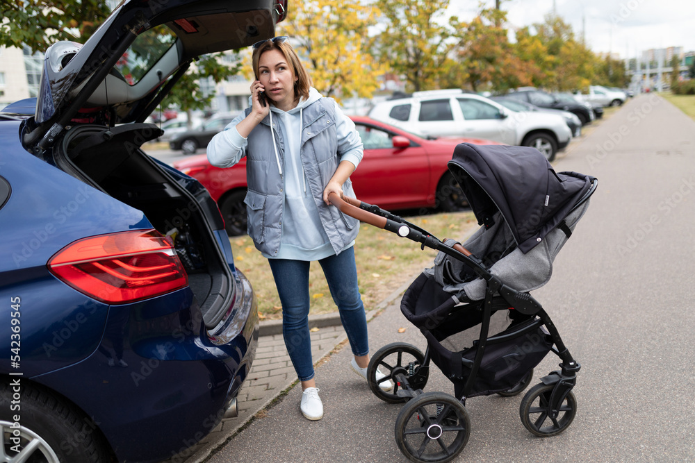 a woman with a baby carriage talking on the phone at the open trunk of a car a woman with a baby carriage talking on the phone at the open trunk of a car