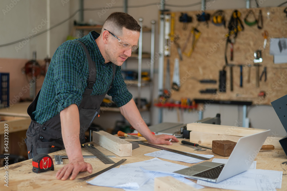 Portrait of a carpenter in protective glasses and work overalls uses a laptop in a workshop. Portrait of a carpenter in protective glasses and work overalls uses a laptop in a workshop.