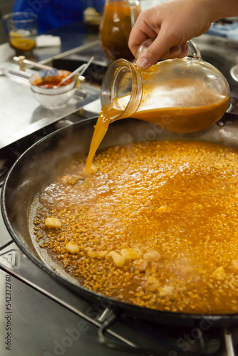 Fototapeta Naklejka Na Ścianę i Meble -  Paella gourmet elaboration stills. A big rice and spices boiling on a paellera while a chef moving hand pours the fish broth to add flavour.