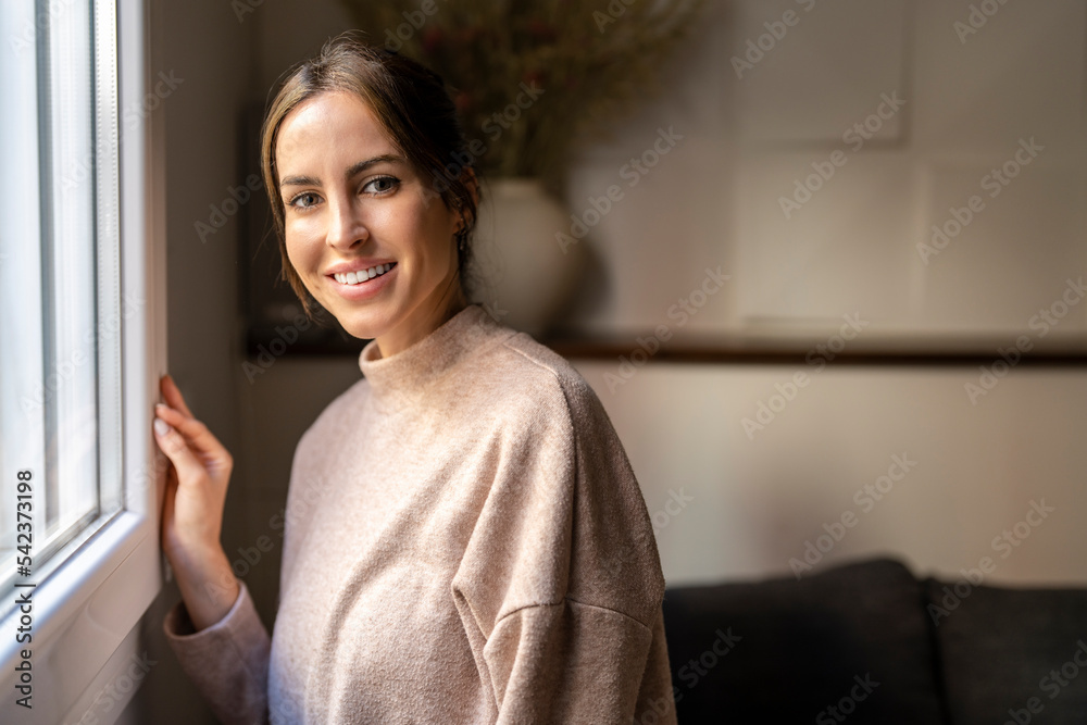 Happy young woman standing by window at home Happy young woman standing by window at home