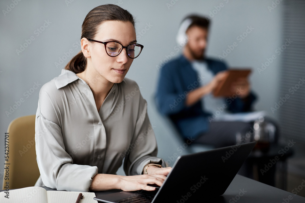 Young serious brunette woman in smart casualwear and eyeglasses sitting in front of laptop and typing while carrying out assignment Young serious brunette woman in smart casualwear and eyeglasses sitting in front of laptop and typing while carrying out assignment
