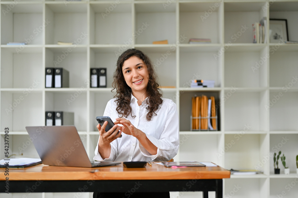Cheerful caucasian businesswoman sitting at her desk, using her phone Cheerful caucasian businesswoman sitting at her desk, using her phone