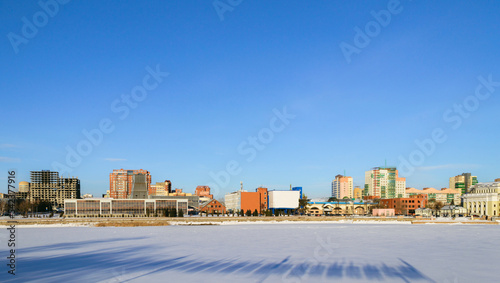 Panorama of a frozen river and buildings illuminated by the sun on a winter day against a blue sky