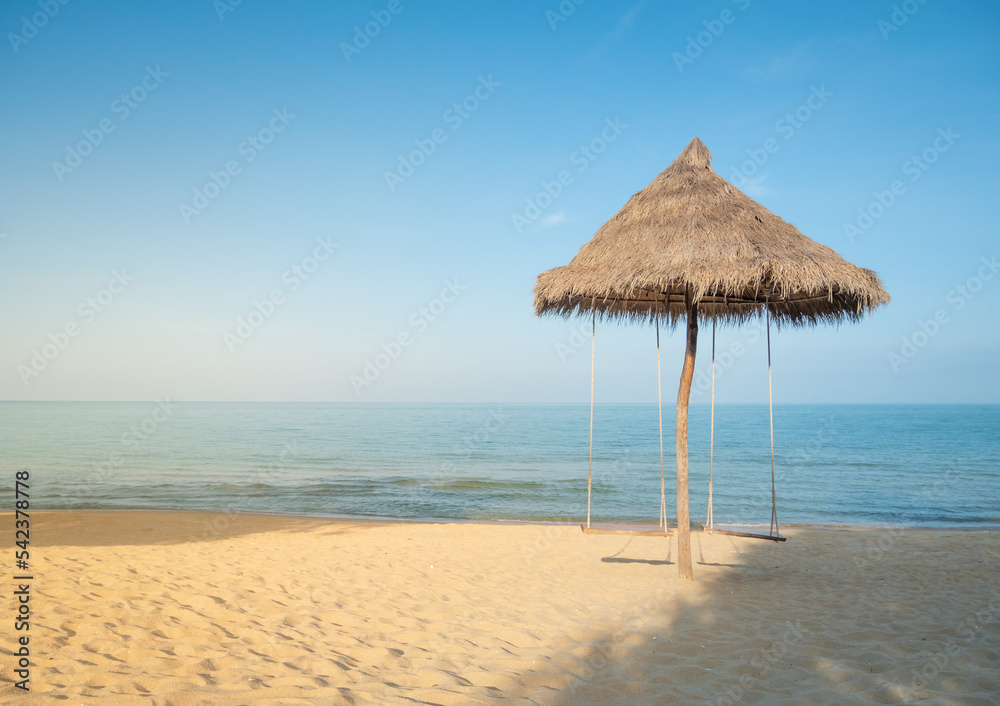 The beach umbrella is made of natural grass with 2 swings hanging. There was a sea and blue sky in front.