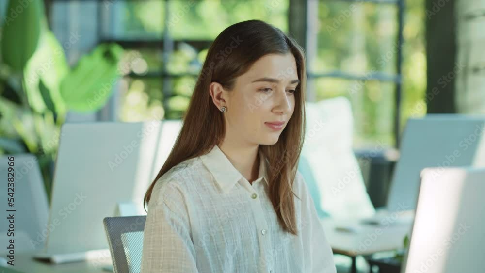 Portrait of a young working woman sitting using a computer looking at a camera smiling feel happy in the light office. Attractive long-haired lady. Modern workspace. Technology. Slow motion