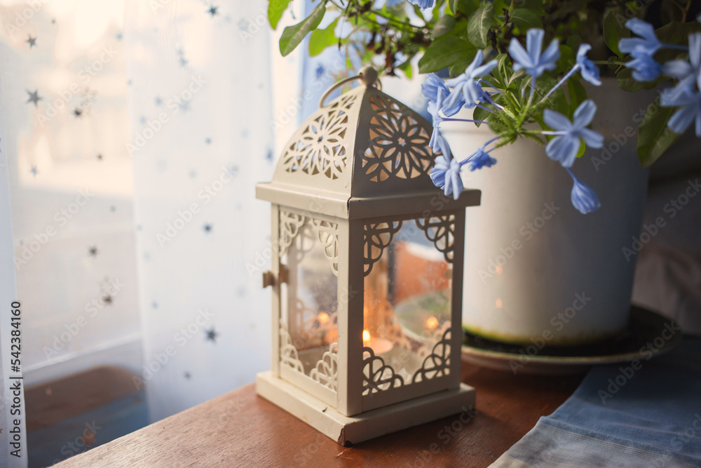 Ornate vintage style lantern with burning candle inside standing near blooming blue flower pot on table near window. Modern new retro Provence style. Summer in countryside.Film grain. Soft focus.