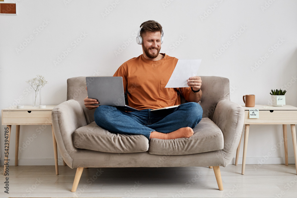Happy young man with laptop on his knees looking at paper with points of new subject while sitting on couch during online lesson