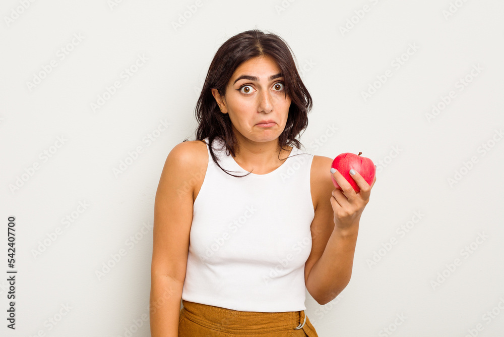 Young Indian woman holding an apple isolated on white background shrugs shoulders and open eyes confused.