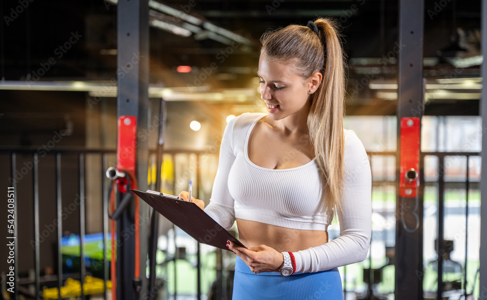 Fototapeta premium Young fitness instructor making schedule for a client on the clipboard at the gym. Attractive blonde trainer writing report for a client.