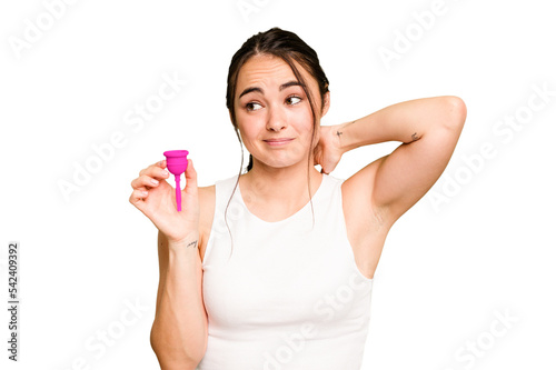 Young caucasian woman holding menstrual cup isolated on green chroma background touching back of head, thinking and making a choice.