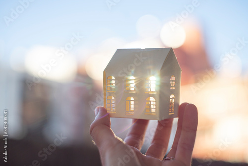 A hand holding a small white paper house illuminated by back sunlight. Beautiful blurred background.