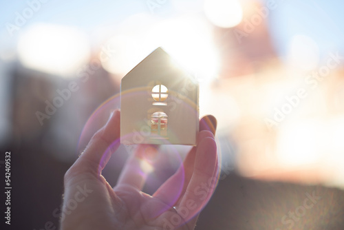 A hand holding a small white paper house illuminated by back sunlight. Beautiful blurred background and Sun flare.