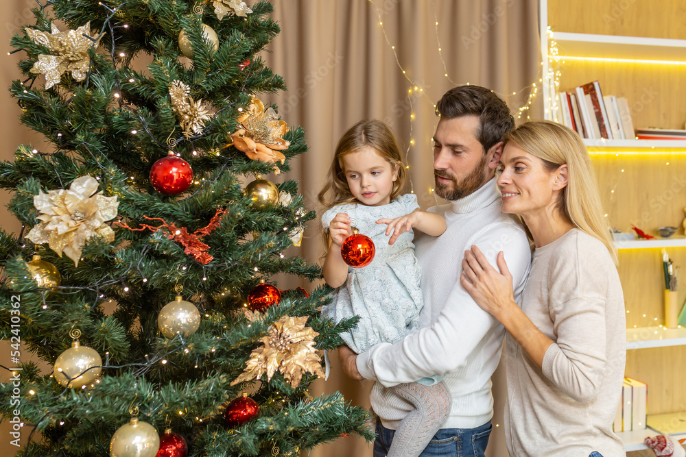 A young happy family decorates a Christmas tree for the Christmas holidays. Dad holds his daughter in his arms, the girl hangs a toy, mom hugs dad. A young happy family decorates a Christmas tree for the Christmas holidays. Dad holds his daughter in his arms, the girl hangs a toy, mom hugs dad.