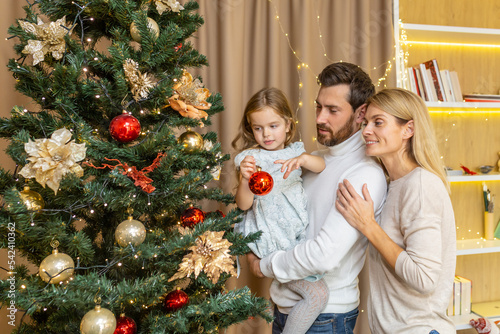 A young happy family decorates a Christmas tree for the Christmas holidays. Dad holds his daughter in his arms, the girl hangs a toy, mom hugs dad.