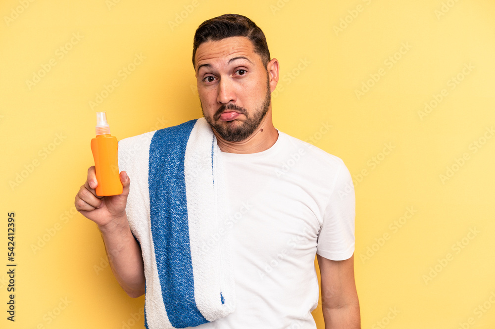Young latin man ready to go to the beach holding a solar skin protection isolated shrugs shoulders and open eyes confused. Young latin man ready to go to the beach holding a solar skin protection isolated shrugs shoulders and open eyes confused.