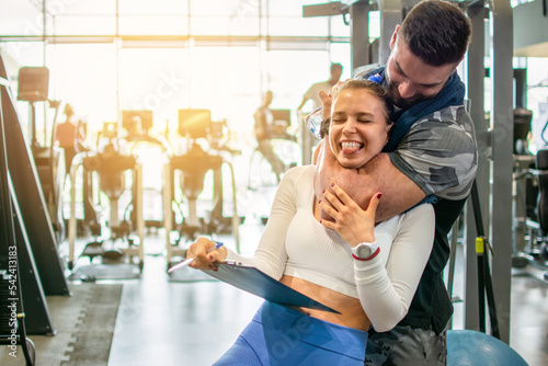 Muscular fit man choking a young female personal trainer with his arm muscle at the gym. Friends having fun at the gym. photo