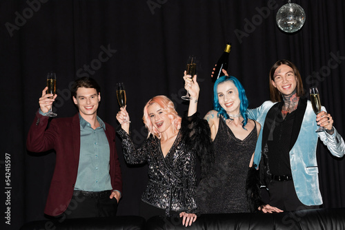happy and stylish queer people toasting with champagne while celebrating christmas and looking at camera on black background