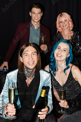 tattooed man with champagne bottle looking at camera while celebrating christmas with nonbinary friends on black background