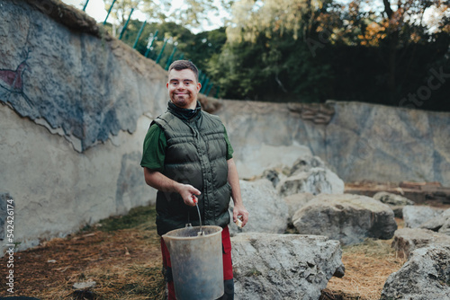 Caretaker with down syndrome in zoo giving food in animal enclosure. Concept of integration people with disabilities into society.