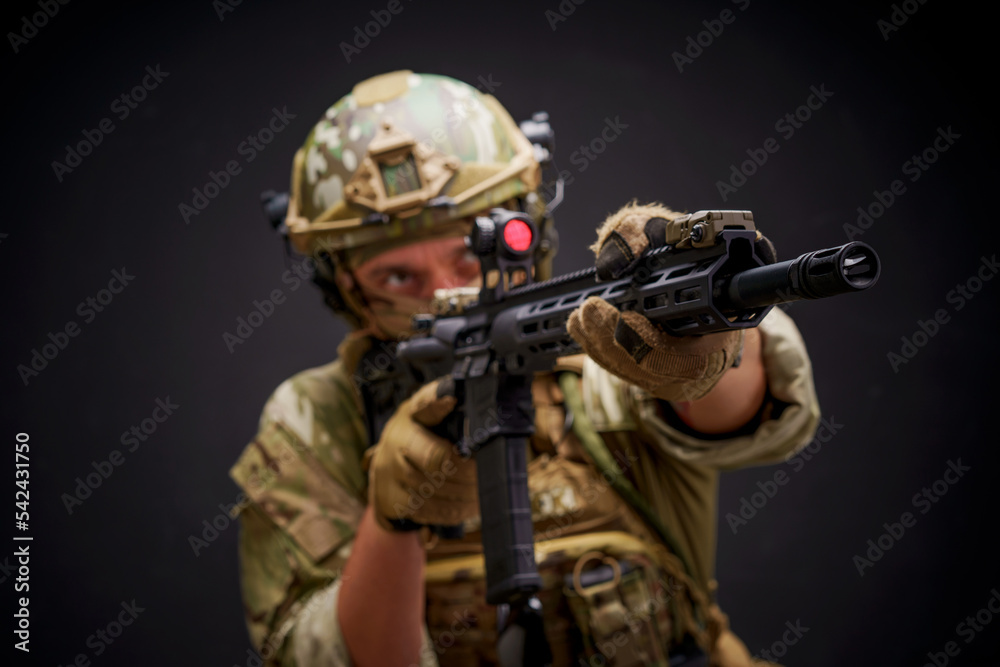 Military man in camouflage holding assault rifle target acquisition through monocular. Special forces man dressed up in military ammunition with gun isolated on black background aiming for a shot