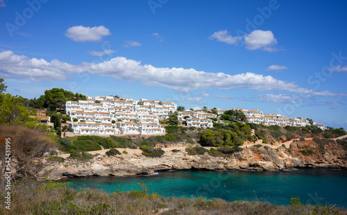 View of the coast of Mallorca in summer. Rocky cliff with turquoise clear water. White mediterranean houses in green bushes. Above blue sky with clouds. Vacation, recreation, tourism