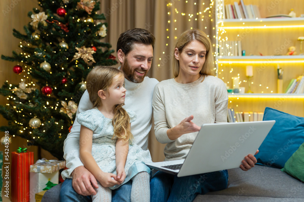 mature parents teach their child to use a laptop studying learning online browsing internet together, sitting on sofa in living room background Christmas tree father and mother with kid daughter. mature parents teach their child to use a laptop studying learning online browsing internet together, sitting on sofa in living room background Christmas tree father and mother with kid daughter.