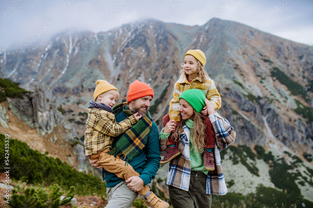 Happy parents with their little kids on piggyback at autumn hike, in the middle of mountains. Concept of healthy lifestyle.
