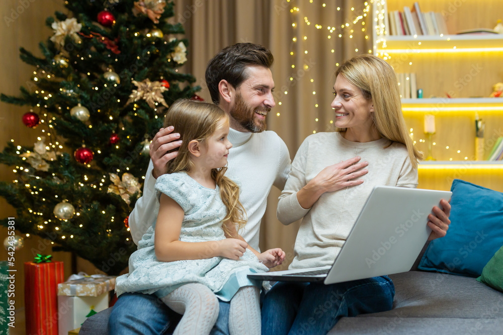 mature parents teach their child to use a laptop studying learning online browsing internet together, sitting on sofa in living room background Christmas tree father and mother with kid daughter. mature parents teach their child to use a laptop studying learning online browsing internet together, sitting on sofa in living room background Christmas tree father and mother with kid daughter.