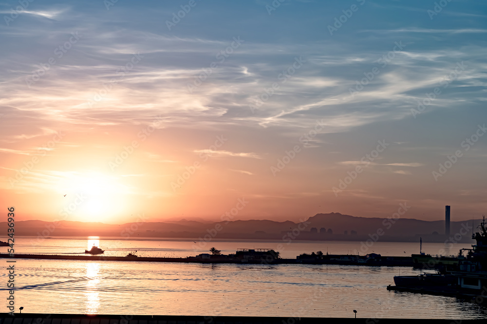 Sunrise at the Port of Algiers city with the silhouette of a trawler sailing in the Mediterranean sea bay, cloudy sky and birds with the great mosque minaret in backlight sunny background. Sunrise at the Port of Algiers city with the silhouette of a trawler sailing in the Mediterranean sea bay, cloudy sky and birds with the great mosque minaret in backlight sunny background.
