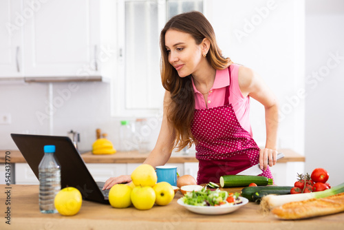 Young housewife using laptop and cooking in home kitchen