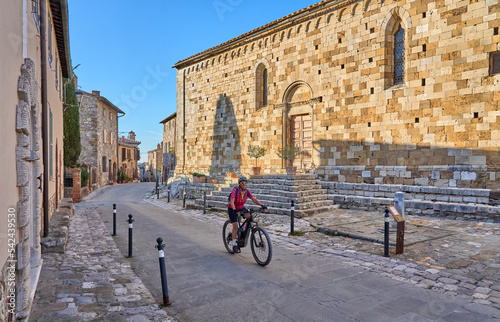 cheerful senior woman cycling with her electric mountain bike in a typical Tuscan village near Pienza, Chianti area,Tuscany, Italy