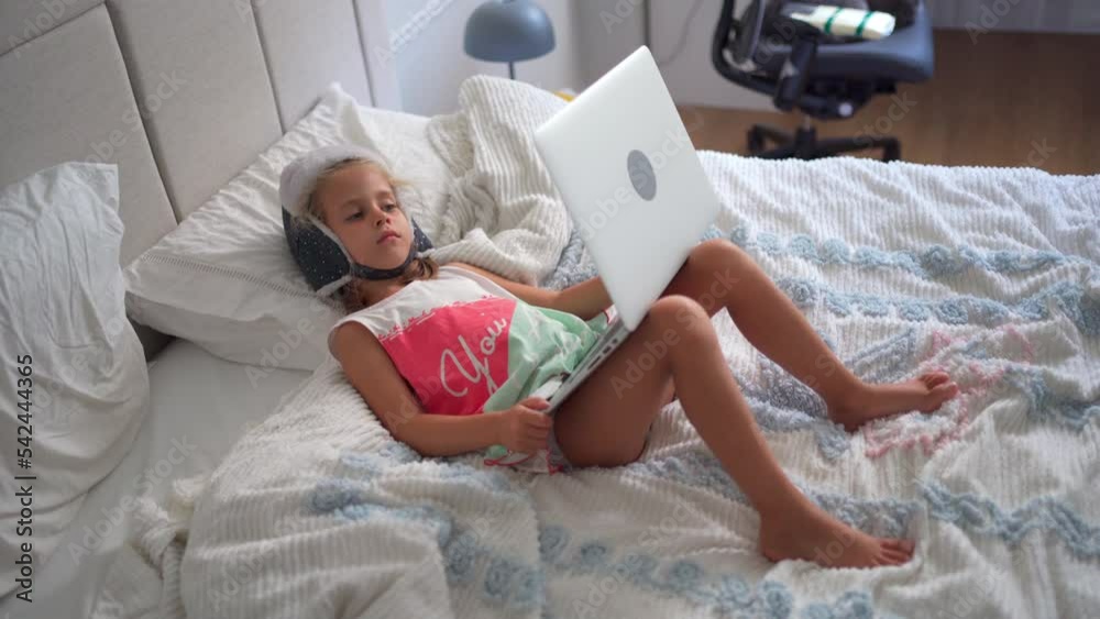 Child in funny hat watching video on laptop lies sofa at home. Little caucasian girl enjoy cartoons on laptop sitting bed in bedroom. Joyful preschool kid girl use laptop computer