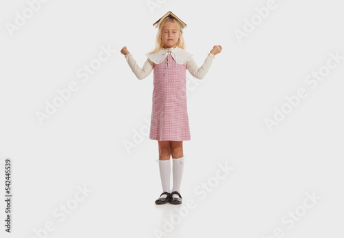 Yoga. Studio shot of little cute stylish girl, kid in retro fashion outfit standing winth book on her head isolated on white background. Back to school concept