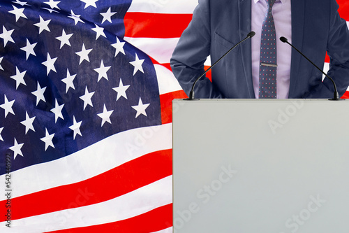 Tribune with microphone and man in suit on US flag background. Businessman and tribune on USA flag background. Politician at the podium with microphones background US flag. Business conference in USA