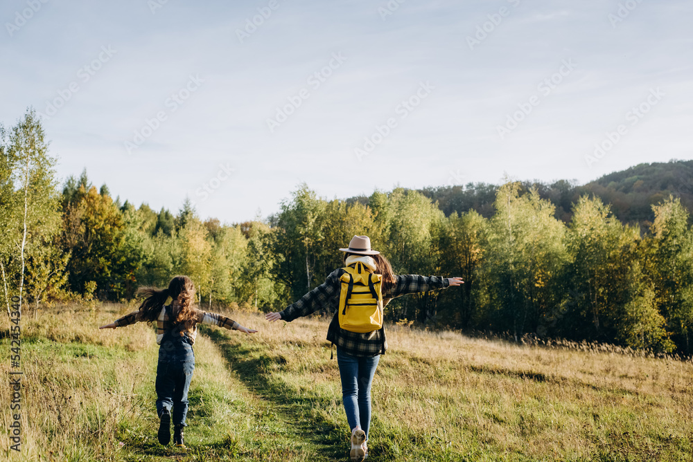 Back view of playful young mother in hat and funny little daughter kid are run grass, playing airplane pilot on background fall forest. Traveler family at sunset day in beautiful autumn park