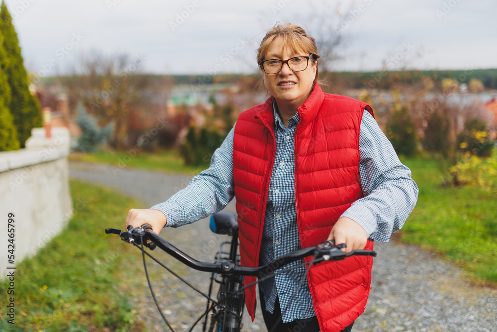 Fototapeta premium Middle aged smiling mature woman holdingt a bike with her hands on the grass on a green field. Summer or Autumn Country Vacation and Adventure Concept