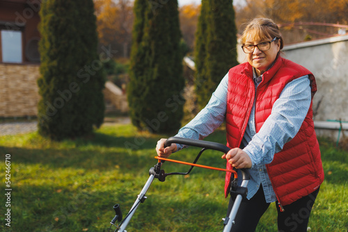 Middle aged woman in red vest using lawn mower on backyard, looking at camera. Female gardener working in summer or autumn, cutting grass in backyard. Concept of gardening, work, nature.