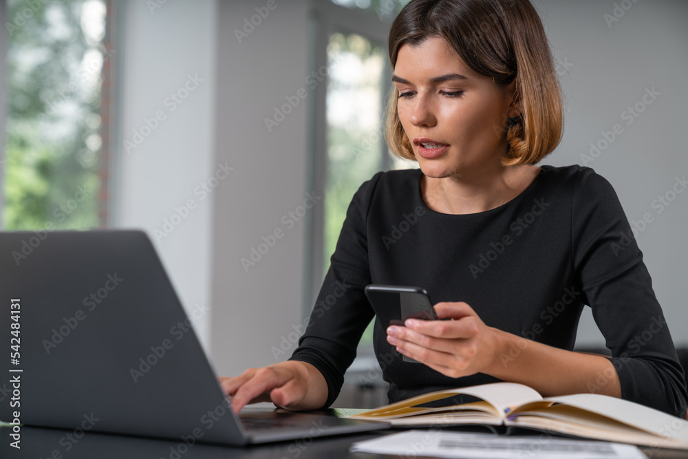 Inspired businesswoman in formal wear typing on laptop holding smartphone Inspired businesswoman in formal wear typing on laptop holding smartphone
