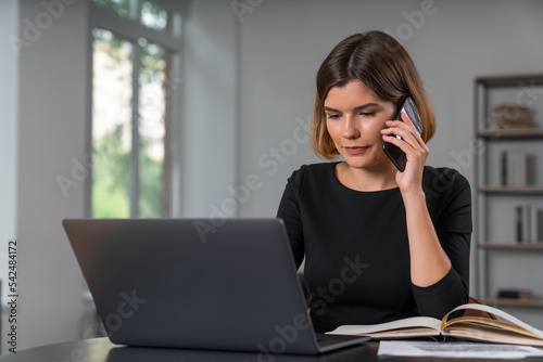 Pleased attractive businesswoman typing on laptop talking on smartphone