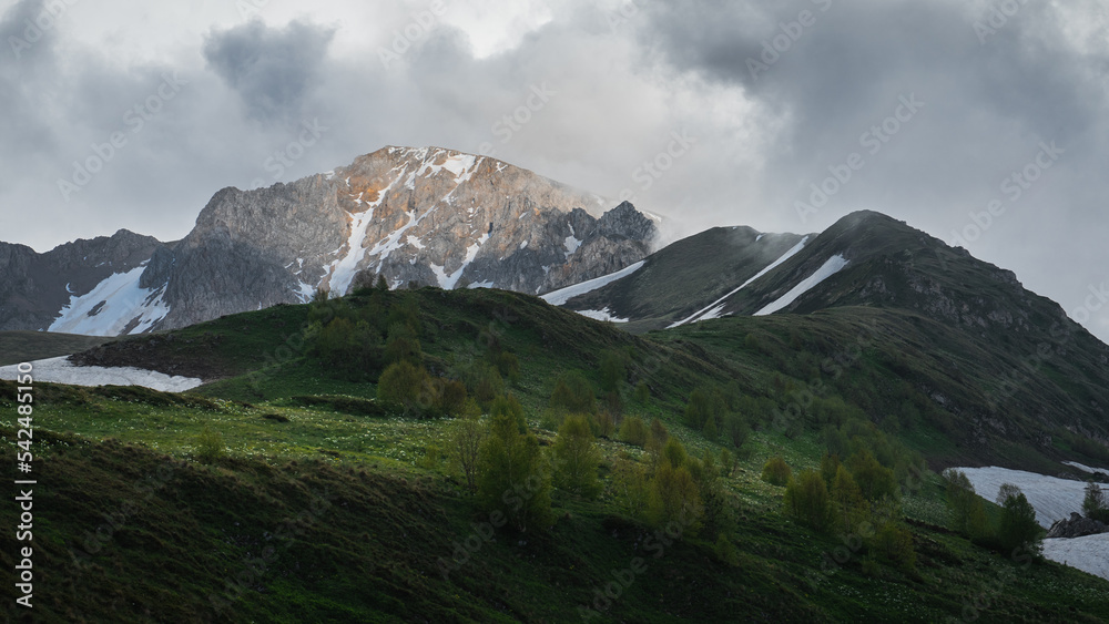 A massive mountain range is covered with clouds, a ray of sun illuminates the mountain and the trees on the hills. A massive mountain range is covered with clouds, a ray of sun illuminates the mountain and the trees on the hills.