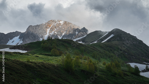 A massive mountain range is covered with clouds, a ray of sun illuminates the mountain and the trees on the hills.