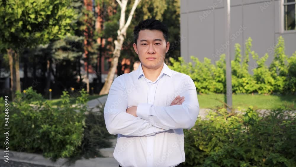 Portrait of young adult serious korean businessman looking at the camera while folding hands outdoor. Successful male business man standing on the background of modern grey glass office center. Portrait of young adult serious korean businessman looking at the camera while folding hands outdoor. Successful male business man standing on the background of modern grey glass office center.