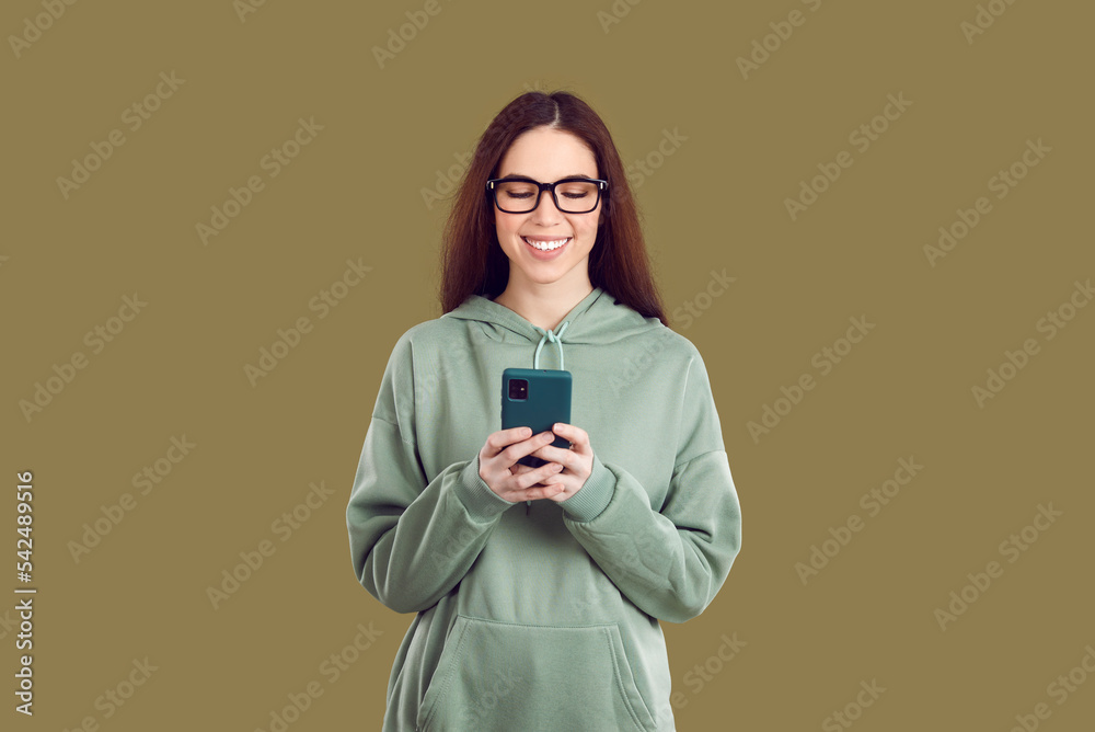 © Studio Romantic - Satisfied pretty young Caucasian woman holding mobile phone in front of using dating apps or social networks to communicate with boyfriend wearing casual sweatshirt stands in green studio