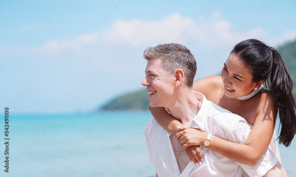 Fototapeta premium Young couple in love outdoor. Happy man giving piggyback ride to his woman and laughing at tropical beach. Girlfriend getting piggyback ride from boyfriend at sunrise in summer.