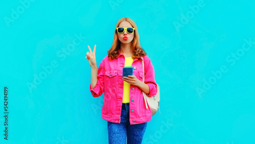 Portrait of stylish young woman blowing her lips sends air kiss with smartphone wearing colorful pink jacket, backpack on blue background