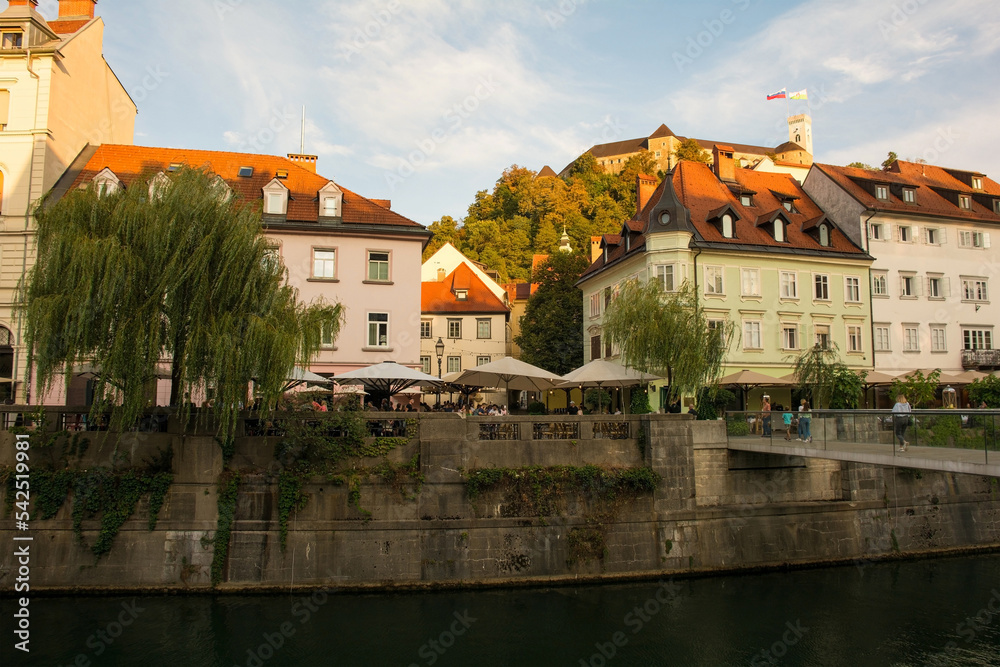 Obraz premium Restaurants and bars on the waterfront of the Ljubljanici River in central Ljubljana, Slovenia. The Fishmarket Footbridge is foreground right, and the castle tower background right