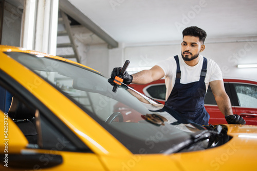 Male bearded worker in blue overalls and protective gloves, on a professional car wash service, applying anti rain coating on a windshield of luxury yellow car. © sofiko14