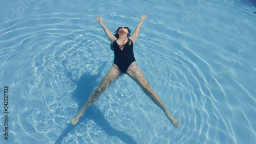 Woman wearing black swimsuit laying on surface of swimming pool as star fish. Girl with white skin floating on water with texture of ripples and sundog outdoors in summer. Spa hotel moments Soft focus