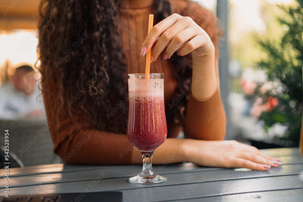 A girl with beautiful curly hair is stirring the freshly squeezed red juice in the cafe.