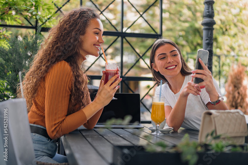 Two girls are sitting in the cafe, looking at something on their smart phone and discussing with a smile.